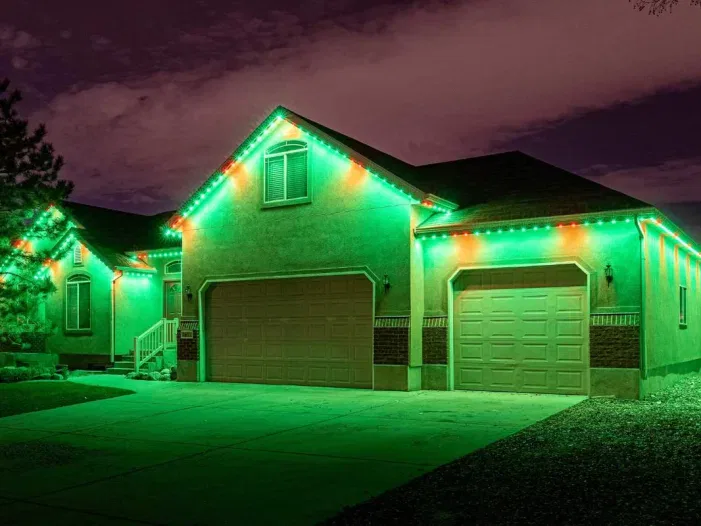 A single-story house at night with green and red lights outlining the roof, garage doors, and front porch. The driveway and yard are illuminated by the colored lights.