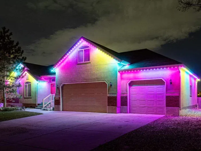 A single-story house at night is lit with bright, colorful LED lights along the roofline, displaying shades of pink, blue, yellow, and green. The driveway and yard are quiet, and the sky is cloudy.
