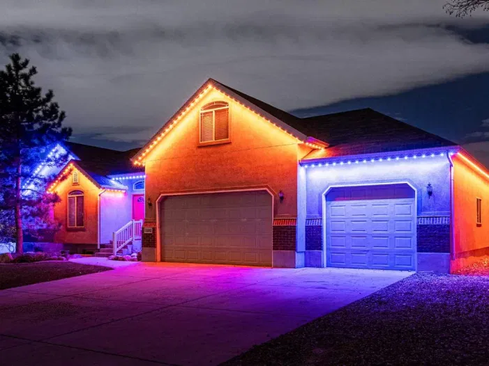A single-story house at night is decorated with red, orange, and blue lights along the roofline and garage, illuminating the driveway and surrounding area.
