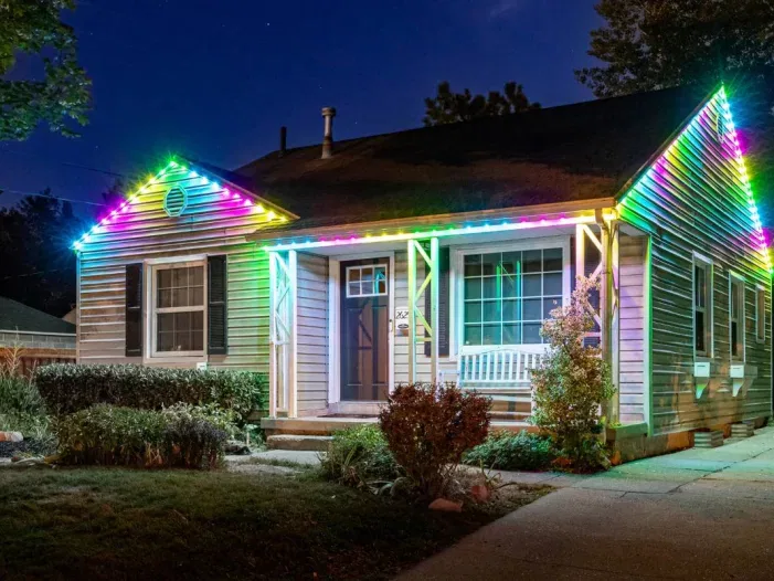 A single-story house at night is decorated with colorful LED lights along the roofline, creating a vibrant, festive glow. Bushes and a white bench sit near the entrance, and the sky is dark blue.