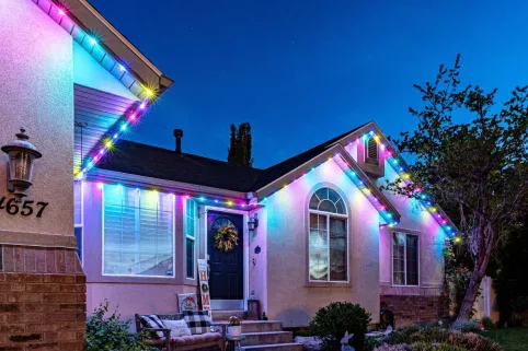 A house at dusk decorated with colorful string lights along the roofline. The lights glow in blue, pink, yellow, and green. A wreath hangs on the front door, and a bench sits on the porch.