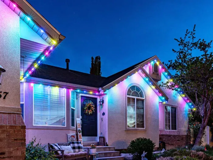 A house at dusk decorated with colorful string lights along the roofline. The lights glow in blue, pink, yellow, and green. A wreath hangs on the front door, and a bench sits on the porch.
