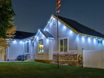 Single-story house at night with white string lights along the roofline, a manicured lawn, a flagpole, a tree on the left, and a white fence on the right. The sky is dark with faint stars visible.
