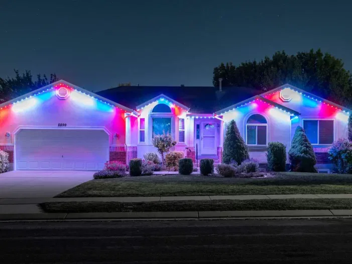 A single-story house is decorated with blue, red, and white lights along the roofline at night, with bushes and trees in the front yard and a driveway leading to a double garage.