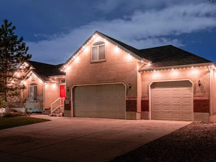 A single-story house at dusk with a glowing red front door, warm exterior lights outlining the roof, three garage doors, a large driveway, and a small tree by the entrance.