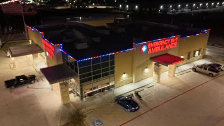 Aerial night view of an emergency medical facility with bright red “EMERGENCY 24/7 AMBULANCE” signs, blue and red lights along the roofline, parked vehicles, and a designated handicap parking space near the entrance.