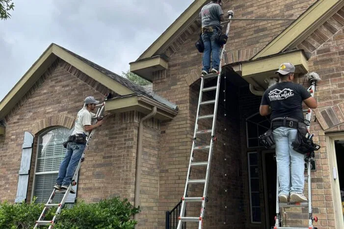 Three workers on ladders install or repair fixtures near the roof of a brick house. The sky is overcast, and greenery surrounds the front porch area.