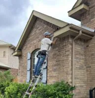 A person wearing a cap and tool belt stands on a ladder, working on the soffit of a brick house next to a bush, with another ladder positioned nearby under a cloudy sky.