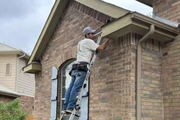 A person wearing a cap and tool belt stands on a ladder, working on the soffit of a brick house next to a bush, with another ladder positioned nearby under a cloudy sky.
