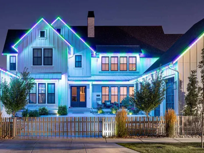 A large modern house at night, outlined with bright blue, green, and purple LED lights along the roof, with a white picket fence, trees, and a well-kept front yard.
