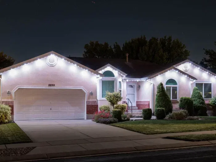 Single-story suburban house at night with white holiday lights outlining the roof, a two-car garage, trimmed bushes, and a well-maintained front lawn and pathway.