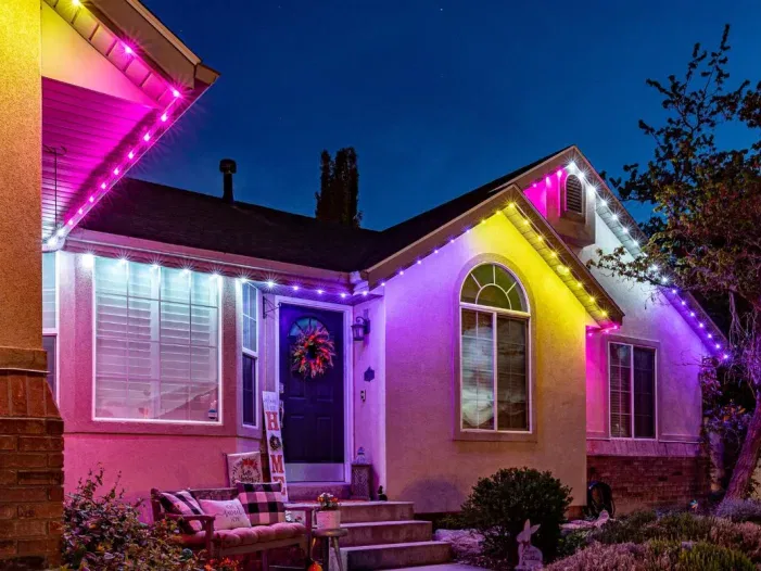 A house at night decorated with bright pink, purple, and white lights along the roofline, illuminating the front porch and windows. There is a wreath on the door and a bench with cushions outside.