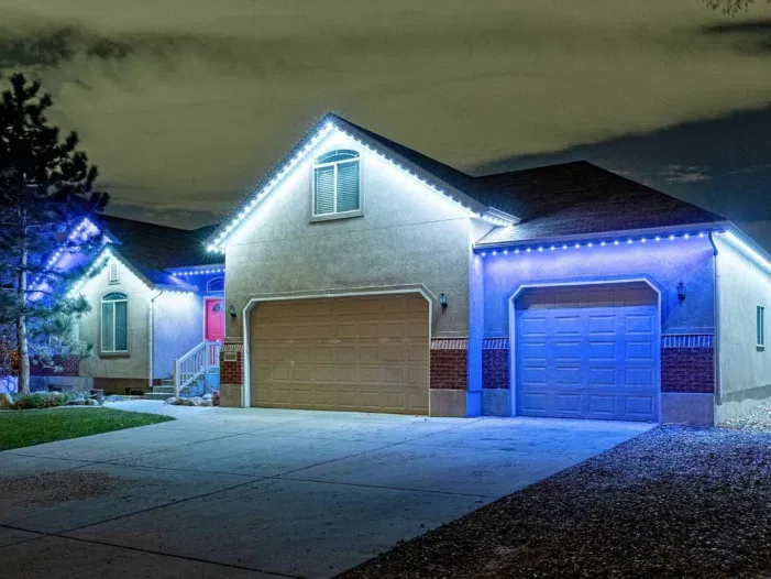 Single-story house with two garages is decorated with bright blue and white lights outlining the roof at night. The driveway is empty, and the yard includes gravel, grass, and a tree.
