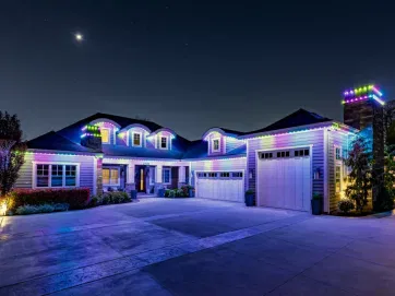 A large house at night is outlined with colorful LED lights along the roof and chimney. The driveway is wide and empty, and a crescent moon is visible in the dark, starry sky above the home.