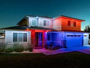 A two-story suburban house is lit with red, white, and blue lights along the roofline at night, creating a festive and patriotic appearance. The garage and front yard are visible under the colorful glow.