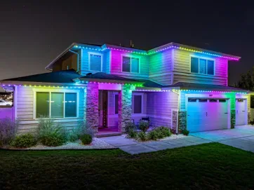 A two-story suburban house at night, outlined with colorful LED lights in blue, green, pink, and purple, illuminating the exterior and creating a vibrant, festive look.