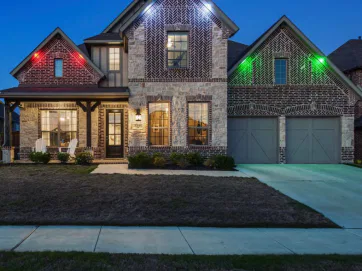 A two-story brick and stone house with front porch chairs, a double garage, and colorful red, green, and blue exterior lights illuminating the facade at dusk.