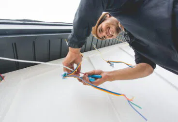 A smiling electrician, viewed from below, installs or repairs colorful electrical wires on the exterior wall of a building. He uses tools and works near a black roof edge against a bright sky.