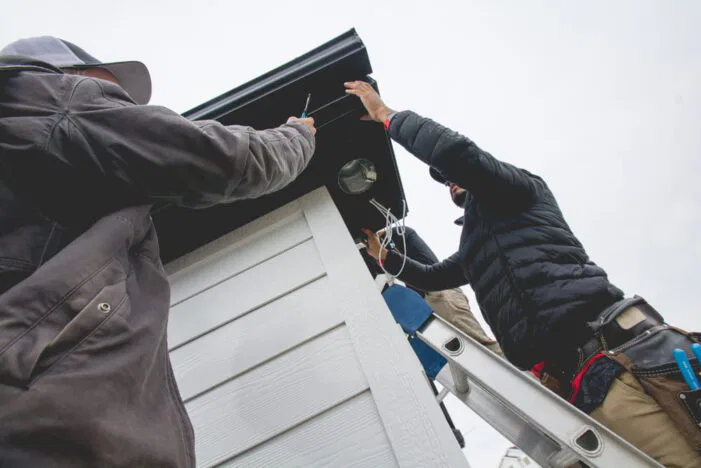 Two people stand on ladders installing or repairing something under the roof eaves of a white building, using tools and wires, with an overcast sky in the background.