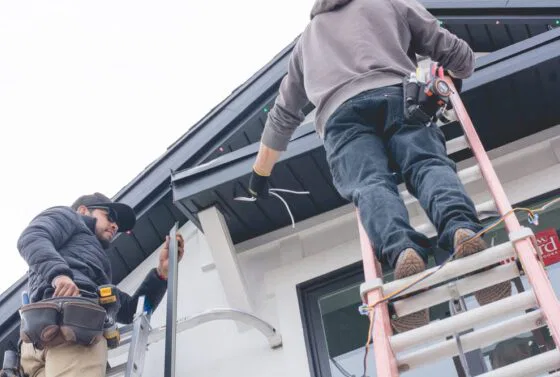 Two workers installing or repairing a roof; one stands on a ladder handling wires, while the other stands on the ground holding a metal piece. Both are wearing work gear and the scene is outside a house.