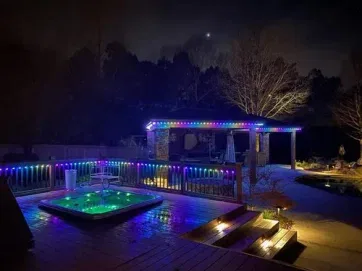 A backyard at night with a lit-up gazebo and hot tub, both outlined by colorful string lights. The deck is wet, and nearby trees and a pool are visible under a dark, partly cloudy sky.