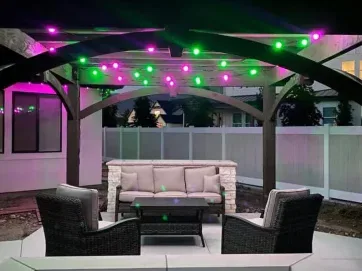 Outdoor patio with a pergola overhead, decorated with purple and green string lights. There’s a small sofa, two chairs, and a table arranged around a fire pit, all set on a paved surface near a white fence at dusk.
