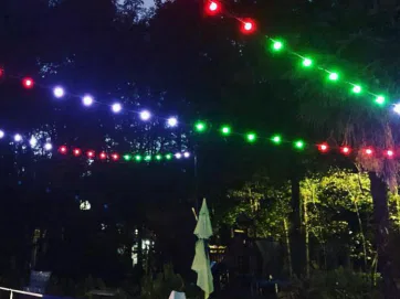 String lights in red, green, and purple hang above a swimming pool at night. Trees and lounge chairs are visible in the background, with an umbrella closed near the poolside.
