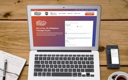 A laptop on a wooden table displays a quote request webpage. Beside it are a notebook with sketches, a pen, a smartphone, and a cup of coffee on a saucer. The scene suggests a workspace setting.