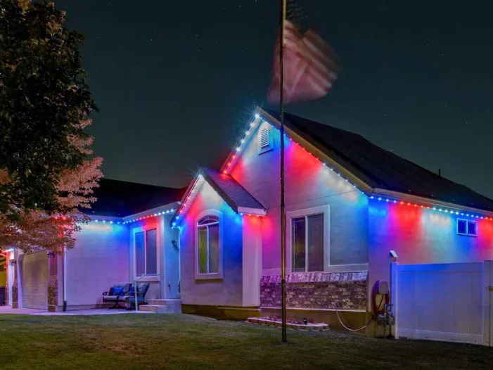 A house at night is decorated with red, white, and blue lights along the roofline. An American flag waves on a pole in the front yard, and a large tree is partially visible on the left.