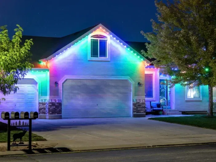 A suburban house at night is decorated with bright, colorful LED lights outlining the roof, illuminating the garage doors and front entrance. Trees, lawn, and mailboxes are visible in the foreground.