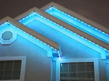A house at night with blue LED lights outlining the roof edges, creating a glowing effect. Two windows are visible below the lit rooflines, and the sky is dark in the background.