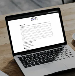 A laptop on a wooden table displays an online request form. Next to the laptop are a cup of coffee on a saucer, a plant pot, and part of a newspaper. The scene appears to be in a casual indoor or outdoor setting.