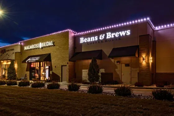 A brick building at night features illuminated signs for “Malmrose Bridal” and “Beans & Brews Coffee House,” with string lights along the roof and small bushes in front.