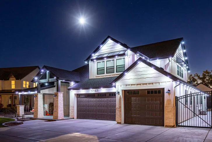 A modern two-story house with white walls, dark roof, and three-car garage, illuminated by bright exterior lights and moonlight at night. A driveway and gated entry are visible in the foreground.