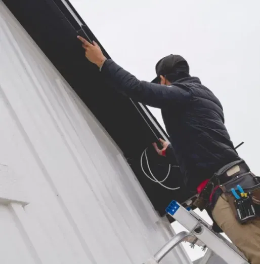 A person on a ladder installs black guttering along the roof edge of a white building, using tools held in a utility belt. The sky is overcast.