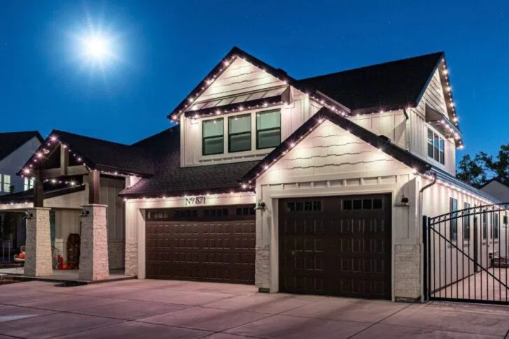 A modern two-story house with white siding and dark garage doors is decorated with string lights along the roofline at night. The full moon shines brightly in the clear sky above.