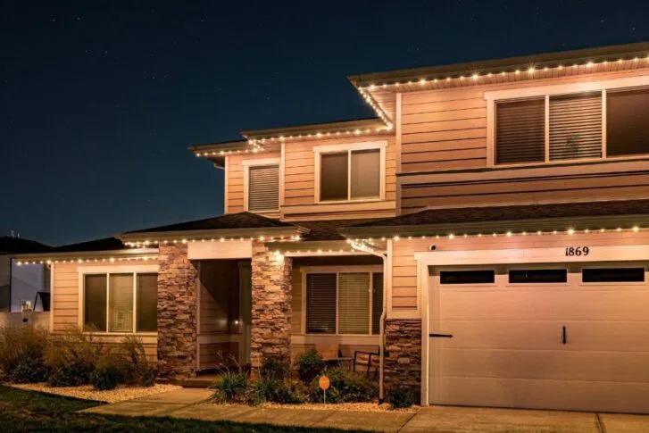 A two-story house with tan siding and stone accents is lit by warm string lights along the roofline at night. The garage door displays the number 1869. The sky is dark with visible stars.