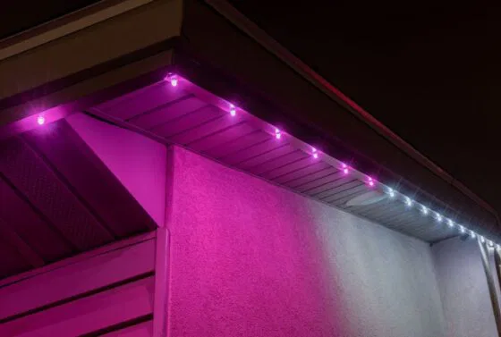 A house roofline at night is decorated with small purple and white LED lights, creating a colorful glow on the exterior walls and eaves.