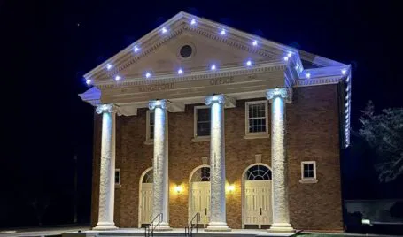 A brick building with four large white columns and three double-door entrances is lit up at night with white lights outlining the roof and columns. The sign above reads Kingsford Office.