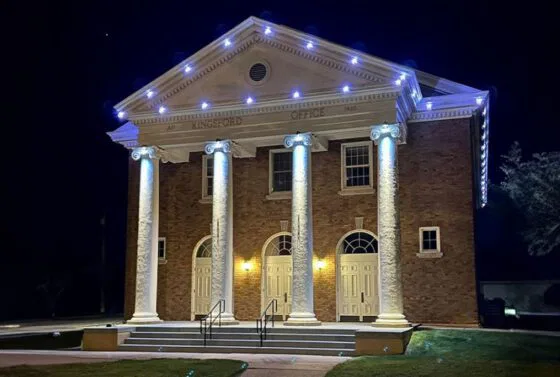 A brick building with four large white columns and three double-door entrances is lit up at night with white lights outlining the roof and columns. The sign above reads Kingsford Office.