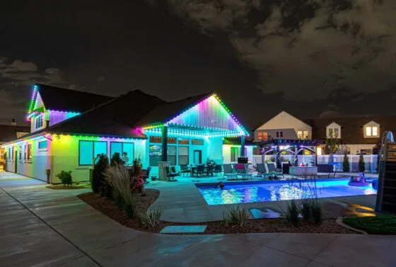 A modern house at night with colorful LED lights outlining the roof, a lit swimming pool in the backyard, and outdoor seating surrounded by landscaping. The sky is partly cloudy.