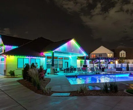 Attached image file A modern house at night with colorful LED lights outlining the roof, a lit swimming pool in the backyard, and outdoor seating surrounded by landscaping. The sky is partly cloudy.