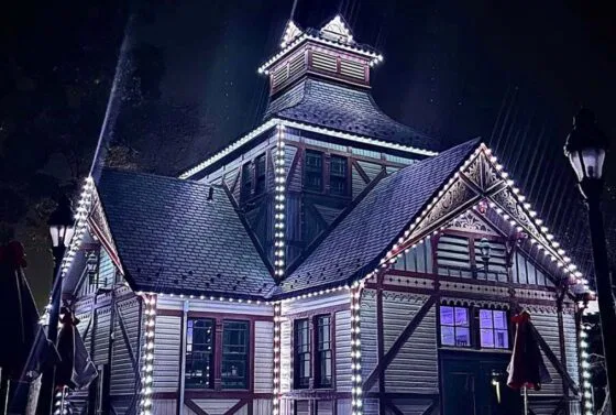A large, ornate building with steep roofs, lit by bright white string lights outlining its edges, stands illuminated at night. The scene has a festive and magical atmosphere.