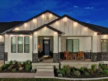 A modern single-story house with white siding, dark trim, and stone accents, warmly lit at night. A small porch features two wooden chairs and a table, surrounded by manicured landscaping and a concrete walkway. House number 5404 is visible.