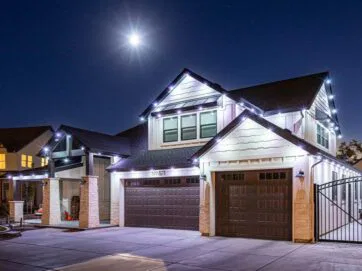 A two-story house at night with bright string lights outlining the roof, three garage doors, and a full moon shining above in a clear sky. The driveway and front yard are visible.