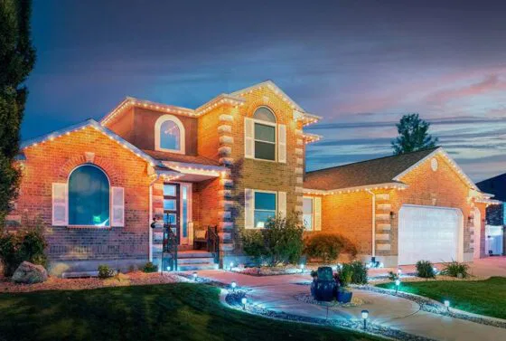 A modern brick house with large windows and decorative lights outlining the roof at dusk. The yard has landscaped plants, a curved walkway, and glowing pathway lights, creating a welcoming atmosphere.