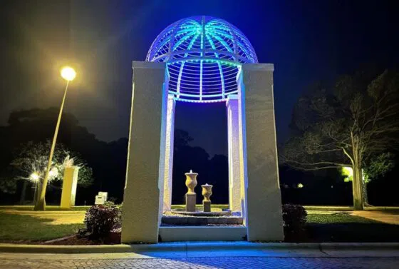 A night scene shows a pavilion-style structure with a domed metal roof lit by blue and white lights, three vases inside, and trees illuminated by lamp posts in the background.
