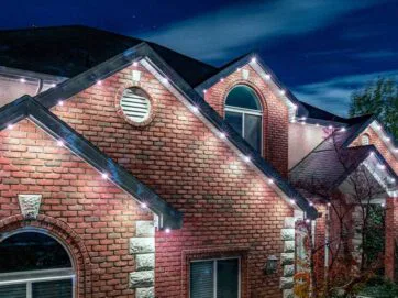 A brick house at night with white exterior lights outlining the roof and gables, creating a festive and welcoming appearance. The sky is dark and clear, and some trees are visible beside the house.