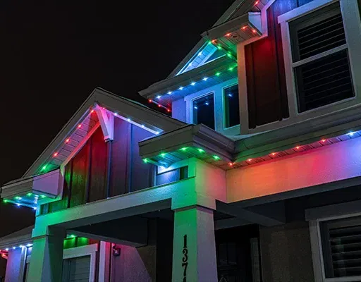 A house at night decorated with multicolored LED lights along the roofline, glowing in blue, red, and green. The lights accent the architectural lines of the home, creating a festive appearance.