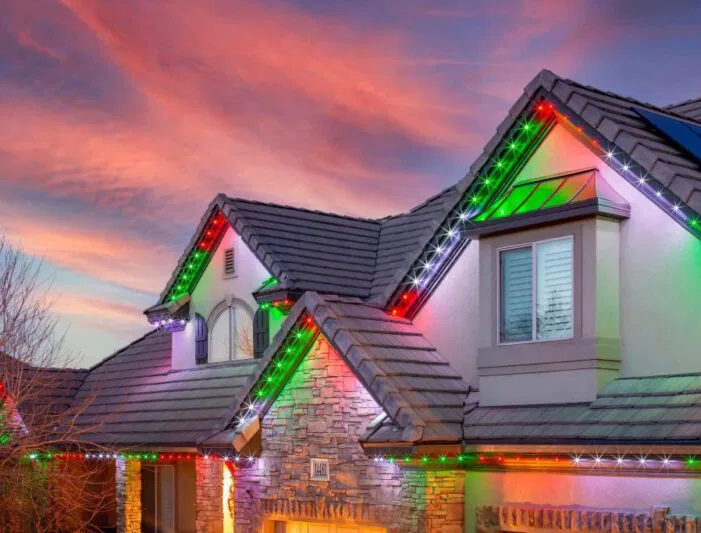 A house with stone accents and a tiled roof is decorated with red, green, and white holiday lights along the roofline. The sky is colorful with dramatic pink and orange clouds at sunset.